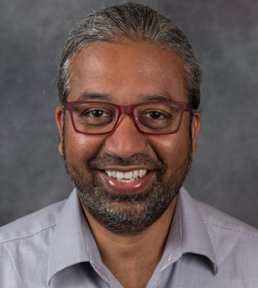 Studio portrait of a smiling man with glasses, gray hair and beard, wearing light shirt.